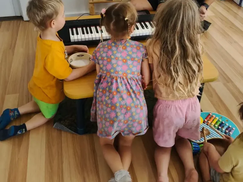 Four young children sit and kneel on a wooden floor around a small table, playing musical instruments including a keyboard, tambourine, and xylophone. An adult, partially visible, sits at the table with them.