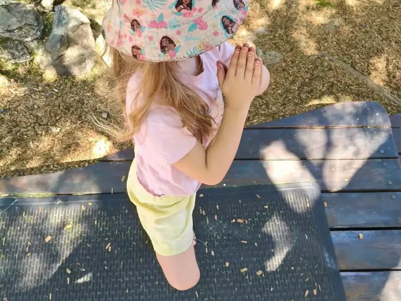A young child wearing a colourful bucket hat, pink shirt, and yellow shorts stands on a mat outdoors with hands clasped, surrounded by sunlight and shadows.