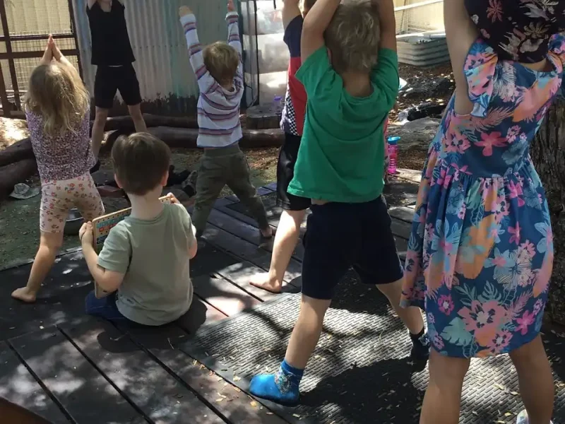 A group of young children follow an instructor outdoors, standing with arms raised above their heads, imitating a yoga or stretching pose under a shaded area near trees and a shed.