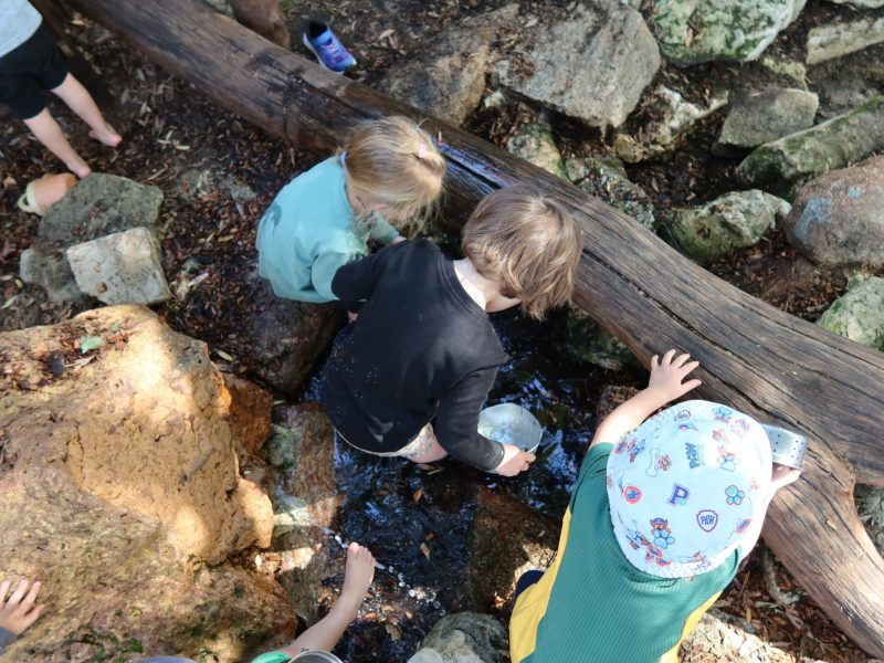 Three Nature Kindergarten children discovering water, rocks and natural materials in the creek bed at Little Learners White Gum Valley Perth